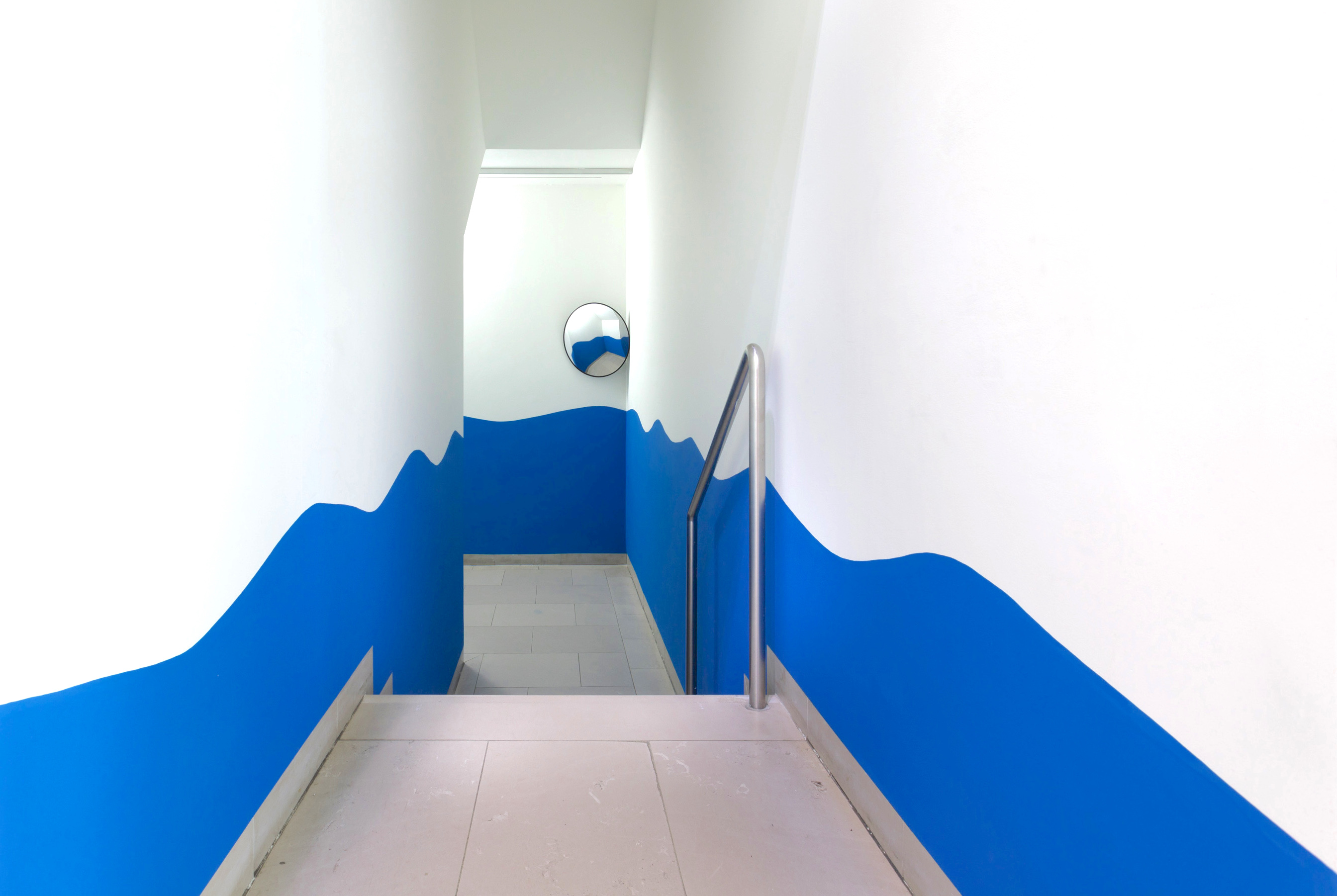 Narrow hallway with stairs and metal railing. The bottom half of the walls 
            are painted blue, ending in a wavy line. A convex mirror is in the right 
            corner of the back wall.