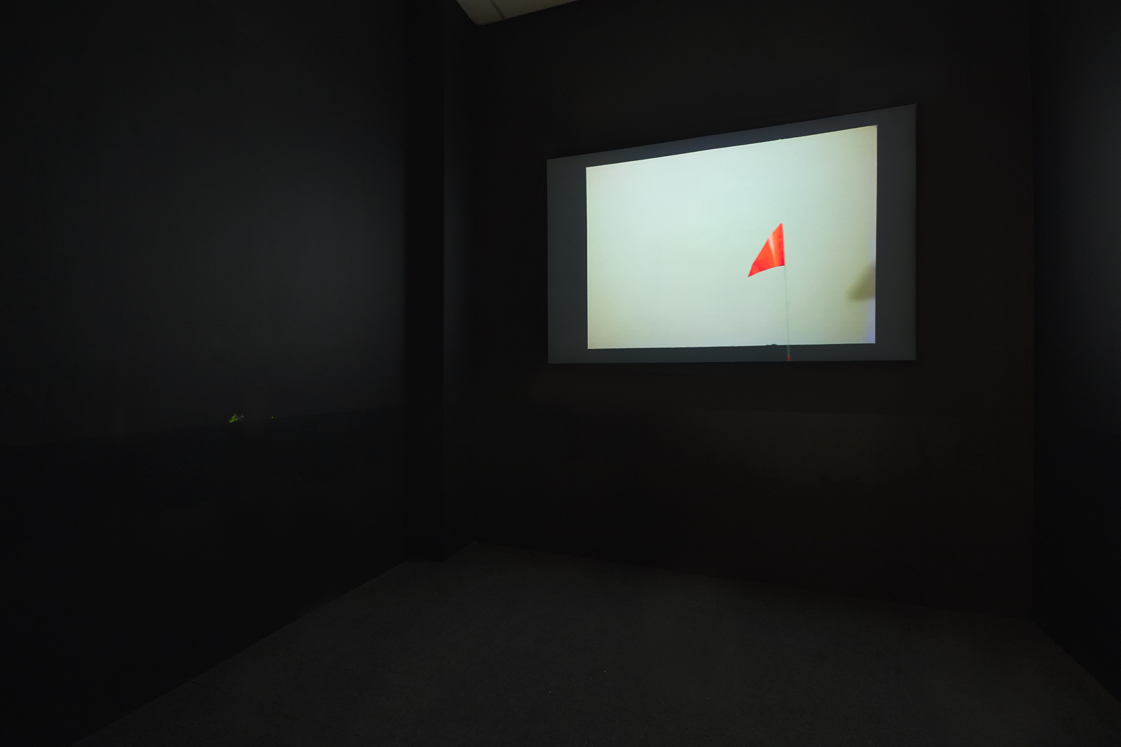A small, dark room painted black, with a projected video on the back wall.
        Video shows a white rectangle with a neon orange pennant flag on a pole in front of it.