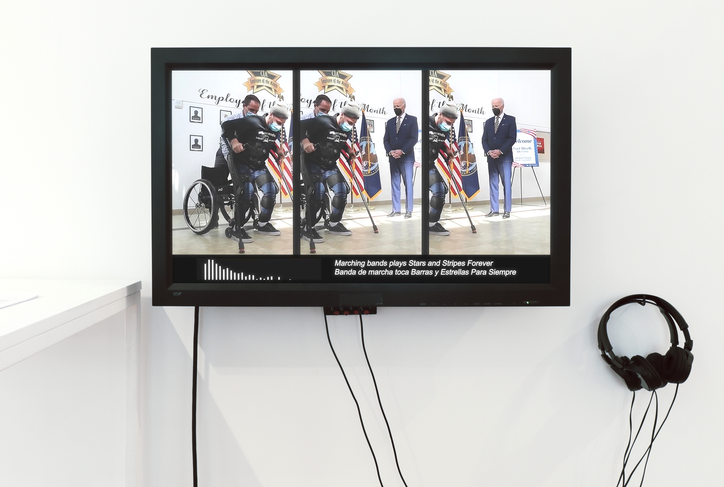 Flatscreen tv with two pairs of headphones hanging on a wall. Video still
            shows person using medical exoskeleton and arm crutches standing
             up from their wheelchair
           while President Biden looks on.