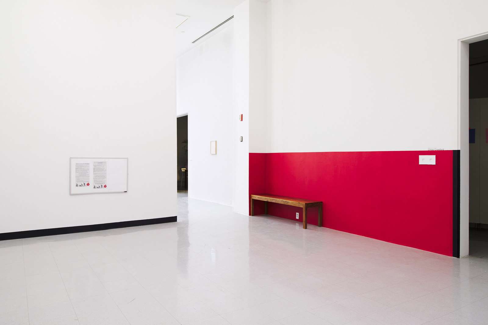 A corner view of the gallery room. There is a doorway in the corner.
        The bottom half of the right wall is painted red with a wooden bench in front of it.
        The bottom half of the left wall is painted a slightly different shade of white as the rest of the walls, and has black trim on the bottom.
        There is a bulletin board on the wall.